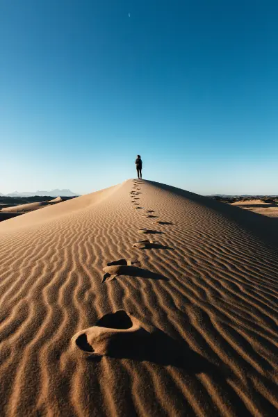 A sand dune with footprints leading up to a woman standing at the peak